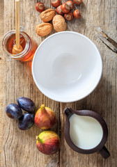 Empty white ceramic bowl, fruits, honey, milk and nuts on wood. Breakfast concept.