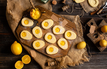 overhead shot of homemade baked egg shaped easter cookies on wooden board on rustic table with yellow eggs, lemon cream. process of baking cookies. easter card