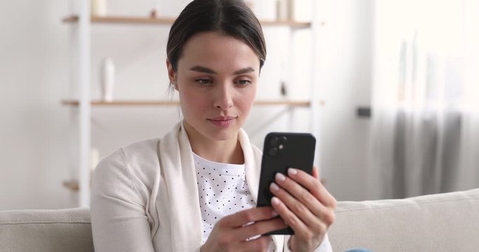 Beautiful young woman looking at smartphone surfing social media dating apps. Pretty millennial girl watching video stories, checking feed, reading news ot texting messages using mobile phone at home.