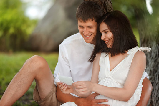 A young man gently hugs his pregnant wife under a tree in nature. A happy couple looks at an ultrasound image of the unborn child.