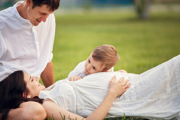 A pregnant girl is lying on the grass, and a little boy is listening to her stomach. Happy family:...