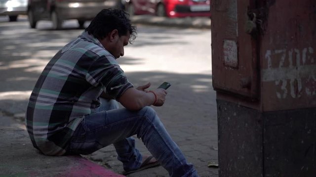 Sad and lonely Indian common man sits on the pavement looking into his smart phone.
