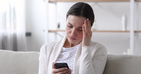 Worried nervous young woman reading bad news in message on smartphone at home. Stressed anxious lady feeling jealous upset holding mobile phone thinking of problem receiving sms sitting alone on sofa. - Powered by Adobe