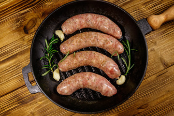 Raw sausages ready for preparation with rosemary, garlic and spices in cast iron grill frying pan on wooden table. Top view