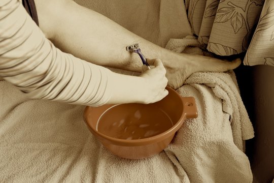 Woman is shaving her leg with long hair in home interior, sepia image.