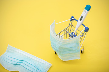 thermometer in a shopping cart and medical masks on a yellow background. Health and vitamin therapy. Protection against viruses and infections. Prevention of diseases transmitted by airborne droplets.