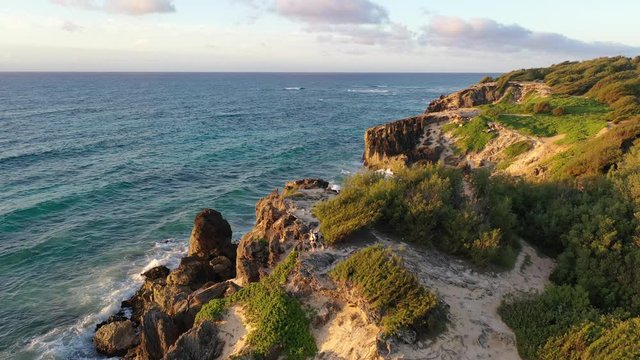 Aerial video of a hiker watching the sunrise from a high lithified sea bluff with limestone and Ironwood pine while waves are breaking on the shore, Makawehi Bluff, Kauai