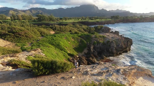 Aerial video of a man hiking left along a rugged sea cliff by the ocean with waves breaking, mountain range in the background and ironwood pine, Makawehi Lithified bluff, Kauai