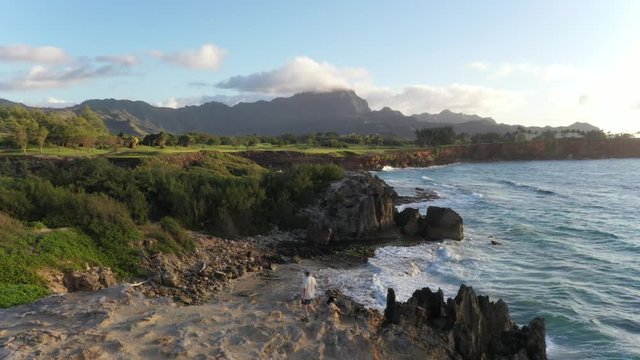 Aerial video of a man walking left along a rugged sea cliff looking at the ocean with waves breaking, mountain range in the background and ironwood pine, Makawehi Lithified bluff, Kauai