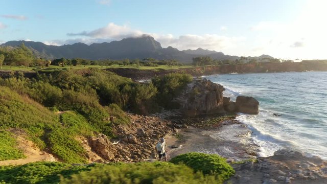Aerial video of a man running right at sunrise along a rugged sea cliff by the ocean with waves breaking, mountain range in the background and ironwood pine, Makawehi Lithified bluff, Kauai