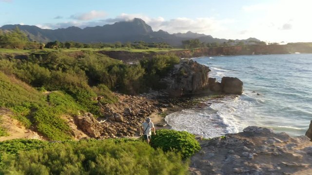 Aerial video of a man running right at sunrise along a rugged sea cliff by the ocean with waves breaking, stopping to look at the view, mountain range in the background and ironwood pine, Makawehi Lit