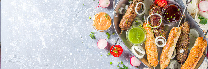 Summer bbq party. Shish kebab on a stick, from ground beef and chicken meat. Lula kebab, traditional Caucasian asian dish. With various sauces, spices and tomatoes, grey stone background, copy space a