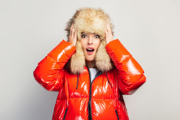 surprised young girl in a red jacket and a fur hat holding hands on his head on a light background....