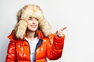 happy young girl in a red jacket and a fur hat, looks away and points a finger at a light...