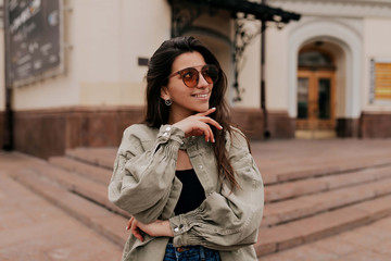 Fototapeta premium Close-up portrait of laughing brunette girl walking on city background. Happy woman in sunglasses and dress walking outdoors. Image of beautiful stylish woman on street on spring day