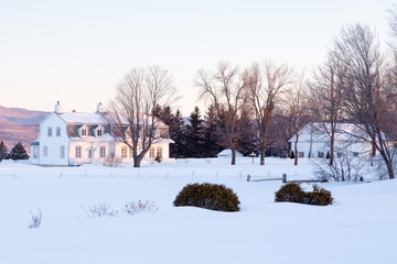 Winter landscape with beautiful patrimonial double white house with Mansard roof and large wooden barn set in snowy field, St. Pierre, Island of Orleans, Quebec, Canada