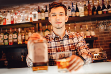 Barman pours whiskey from bottle into glass of ice bar. Dark brown background
