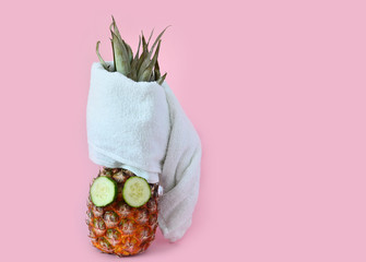 Creative spa concept. Pineapple with cucumbers in the form of an eye mask and a towel on a pink bed...