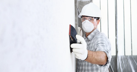 man sand the wall with sandpaper, professional construction worker with mask, safety hard hat,...