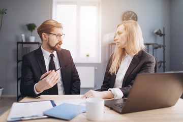Group of two business partners sitting at modern office with laptop and documents and looking at each other during conversation. Competent colleagues discussing new project at work