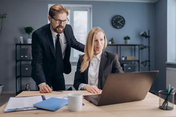 Mature lady with blond hair sitting at working place and looking at computer screen while male business partner standing near and consulting her about new project. Concept of teamwork.