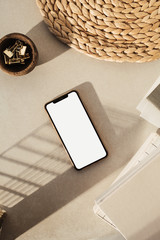 Flatlay of blank screen smart phone, notebooks, clips in wooden bowl, straw stand on beige concrete...