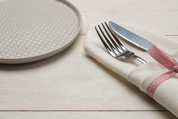 Table setting. plate and cutlery in a white napkin with a pink ribbon, fork and knife on a white wooden table.