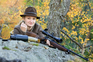 Young, Clever Woman Deer Hunter wearing green uniform with Rifle on the rock in Background autumn...