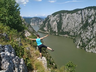 Girl standing on a rocky cliff hundred meters above the Danube Valley between Romania and Serbia, Yoga posing in the sun, meditating with arms wide open 