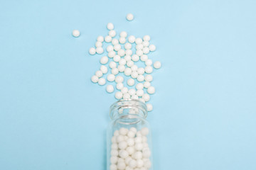 Homeopathic globules scattered from a clear glass bottle on light blue background, alternative homeopathy medicine concept
