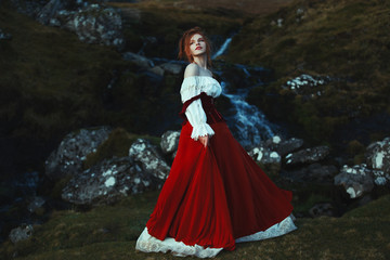 Fototapeta premium Beautiful red-haired girl dressed white and red costume is smiling on the background of stones covered with moss and waterfall