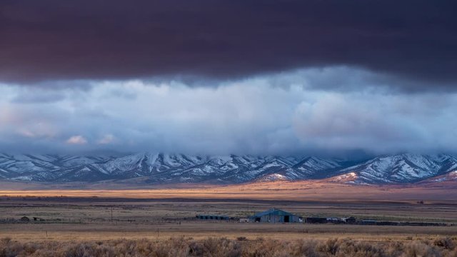 Time lapse of farm in valley with dramatic clouds rolling over mountains covered in snow in springtime in Utah.