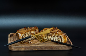 Side view of a cut cinnamon roll with caramel and nuts on a wooden tray with a fork and knife on a black background