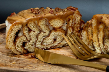 Side view of a cut cinnamon roll with caramel and nuts on a wooden tray with a fork and knife on a black background