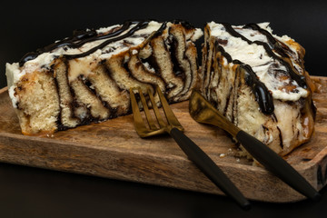 A cut cinnamon roll with white cream and chocolate lies on a wooden tray with a fork and knife on a black background