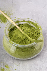 Matcha green tea powder, in a glass container, with a special wooden spatula, gray background, vertical format.