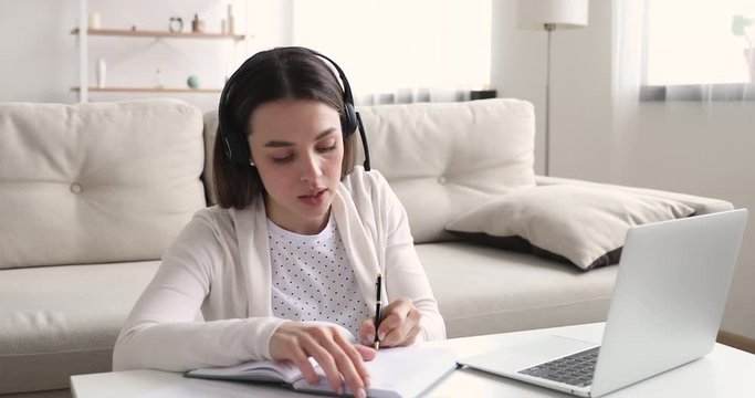 Young girl student wearing wireless headset e learning by web cam chat at home. Millennial woman video conference calling on laptop computer making notes talking by webcam. Distance education concept