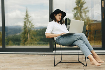 Stylish hipster girl sitting with laptop on terrace at big window. Young happy woman in hat using laptop, shopping or working online from home outdoors. Freelance and freelancer