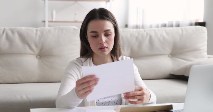 Upset worried young woman reading bad news in paper mail letter. Frustrated girl student opening envelope receiving bank debt notice, high tax bills or failed exam test results concept at home.
