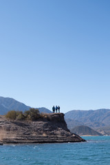 Fototapeta na wymiar three friends gathered on the top of a rock looking at the mountainous landscape at the foot of a lake of calm water with a clear, blue sky