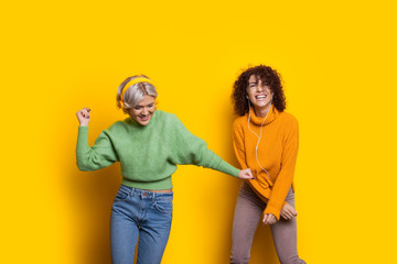 Two beautiful curly haired caucasian women dancing on a yellow background while listening to music from headphones