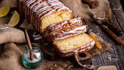 homemade rectangular sliced pound lemon cake with white glaze on top on wooden board on rustic table with sackcloth, lemon slices, vintage forks