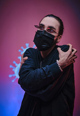 Young caucasian man wearing medical mask, sunglasses and black clothes, standing with hands crossed over coronavirus projection. Isolated in a bright studio, vertical photo