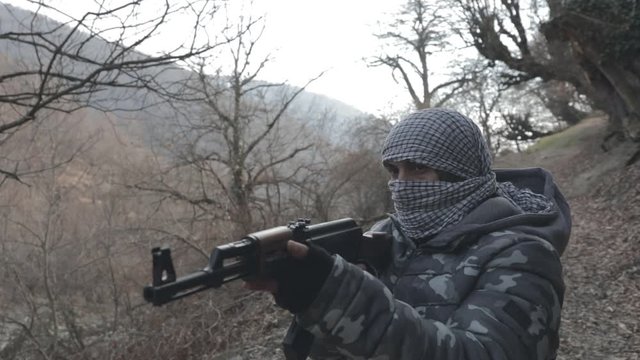 Man in mask with assault rifle ready to attack in the forest. Dangerous bandit aiming gun. Special soldier during the military operation on mountain road. Selective focus