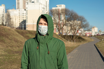 portrait of a guy in a green jacket in a hood in the city. on the face a medical white mask to...