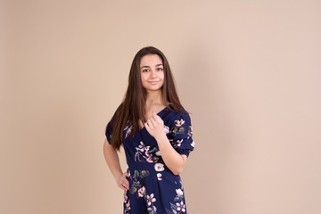 Studio portrait of a girl on a beige background. The girl is looking forward. One arm at the waist, the second pressed to the chest. Playful look. There is a smile on her face. Dressed in a blue dress