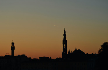 Church towers and the tower of the Palazzo Vecchio, Florence, are seen in silhouette against the colours of sunset sunset. The sky is clear, with a vapour trail left by a plane.