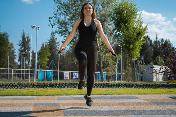 Mujer fitness con ropa oscura brincando la cuerda durante un día soleado con cielo azul 