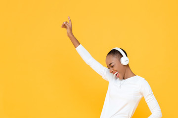 Happy young African American woman raising hand up while listening to music in wireless headphones...