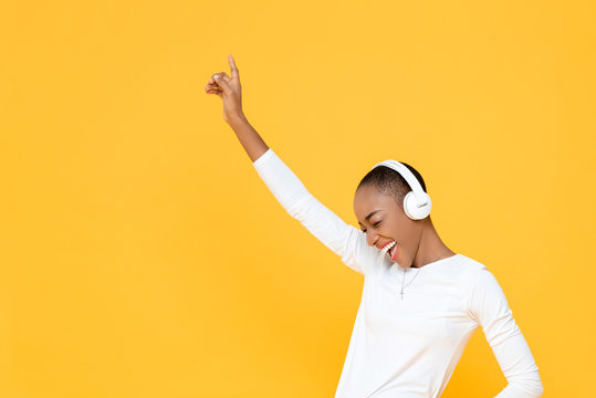 Happy young African American woman raising hand up while listening to music in wireless headphones on yellow isolated studio background